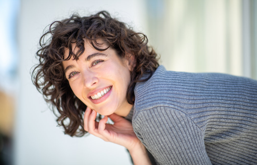 Person with curly hair and a gray sweater smiling and leaning slightly forward against a light background. Atlanta, GA